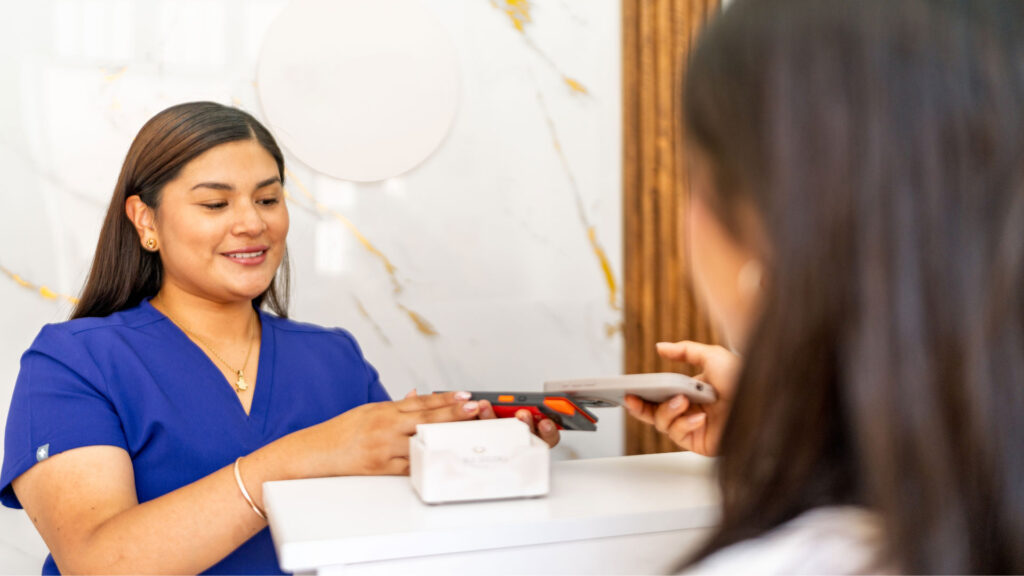 A patient using a mobile phone for contactless payment at a modern dental reception desk with a smiling staff member, illustrating MINISH veneer cost and easy payment options.