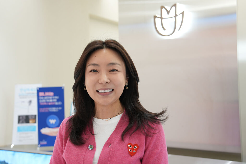 A smiling dental professional in a pink cardigan standing in front of the MINISH Dental Hospital logo in a modern clinic lobby.