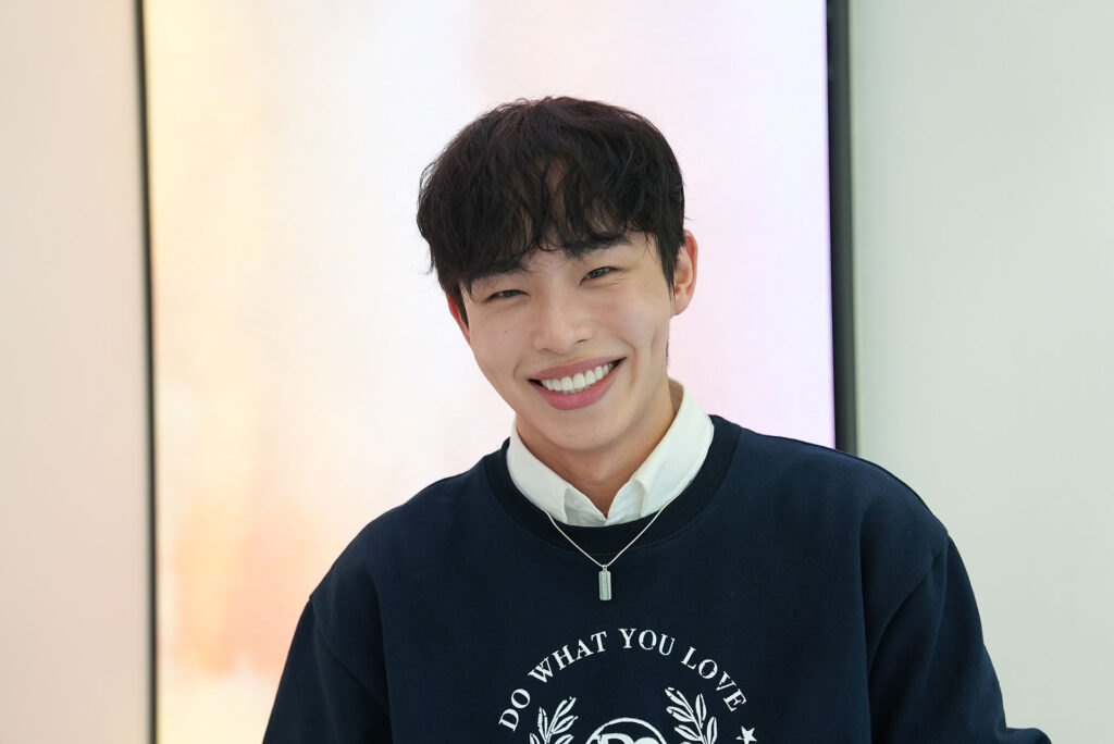 A happy young man with a bright, perfect smile after a MINISH Full Smile Makeover, wearing a navy blue sweater and posing in a modern dental office.